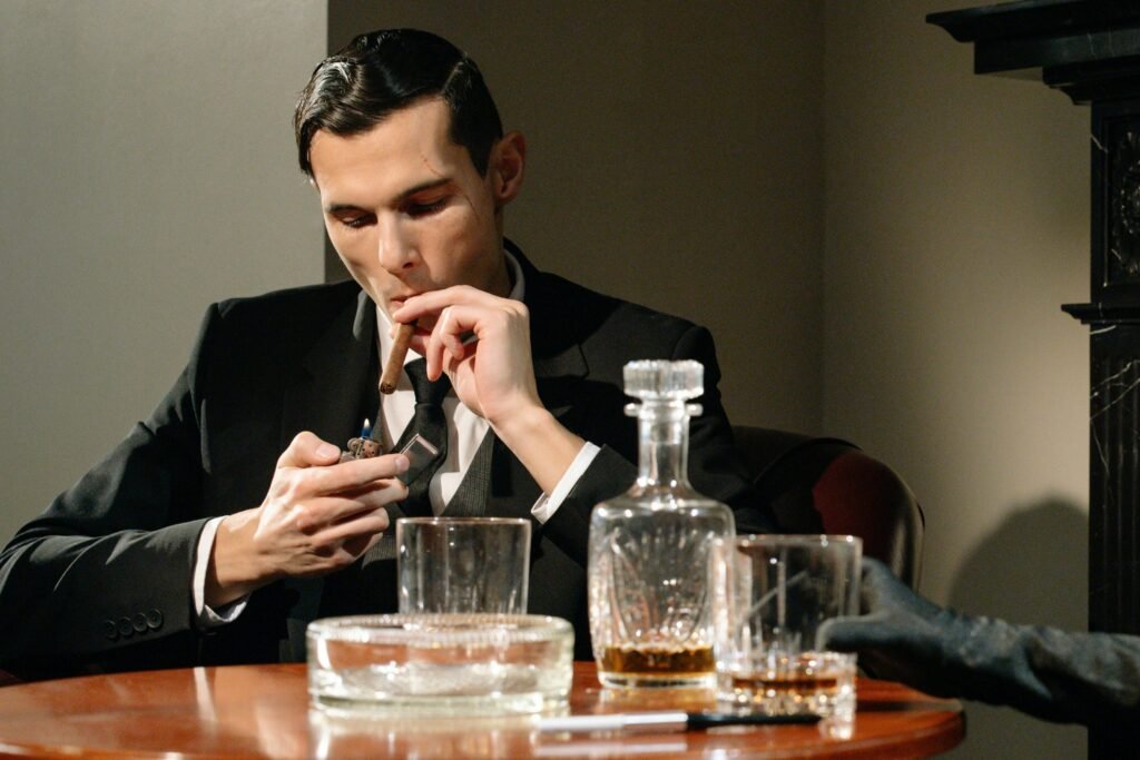 A sophisticated man in a suit smokes a cigar while enjoying a drink, exuding luxury and vintage style.